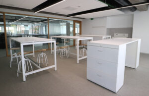 Aula con mesas altas blancas, taburetes y archivador blanco de cajones
