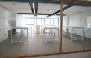 Sala con mesas altas blancas y taburetes de asiento de madera tras mampara de vidrio