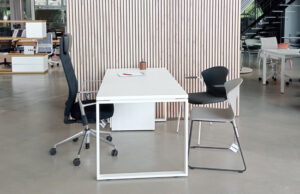 Mesa blanca con silla de dirección y dos sillas de visita ante un panel de lamas