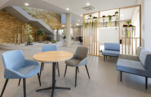 Sala de espera con butacas azules y grises, mesa redonda y muro de piedra con vegetación