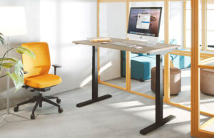 Mesa elevable con tablero gris, silla naranja y mampara acristalada de marco dorado