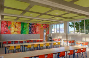 Comedor escolar con plafones acústicos verdes en el techo y mesas con sillas de colores