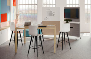 Aula con mesa alta de madera, taburetes blancos y pizarra móvil con pantalla