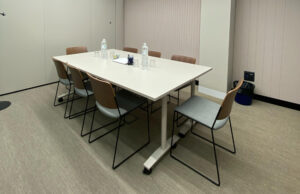 Sala de reuniones con mesa blanca sobre ruedas y sillas de asiento gris y respaldo de cuero