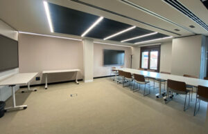 Sala de formación con techo acústico oscuro con luminarias lineales, mesas blancas y pantalla