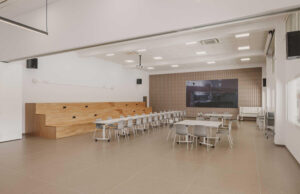 Sala polivalente con grada de madera, mesas plegables, sillas y pantalla