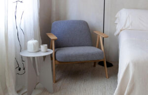 Butaca de madera tapizada en gris junto a una mesa auxiliar blanca con juego de té, al lado de una cama